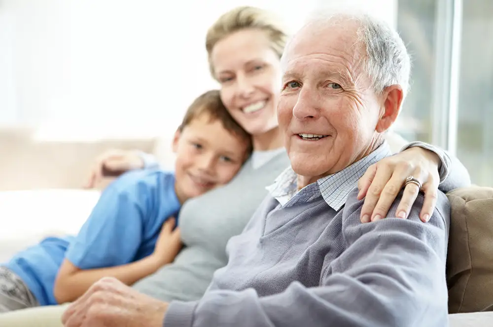 A senior at Village Shalom smiles warmly on a sofa while leaning against his daughter and young grandson, enjoying quality family connection in a supportive memory care community environment.