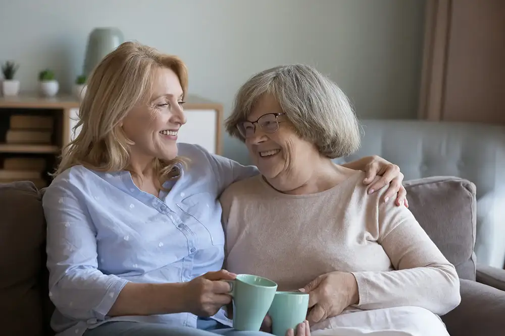 A Village Shalom resident shares a joyful smile and a cup of coffee on a couch with a loved one in a comfortable memory care setting.