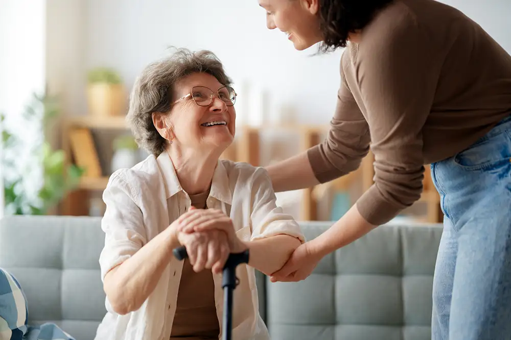 A smiling senior at Village Shalom holding a walking cane receives compassionate assistance from a caregiver, highlighting individualized support for seniors experiencing memory loss.