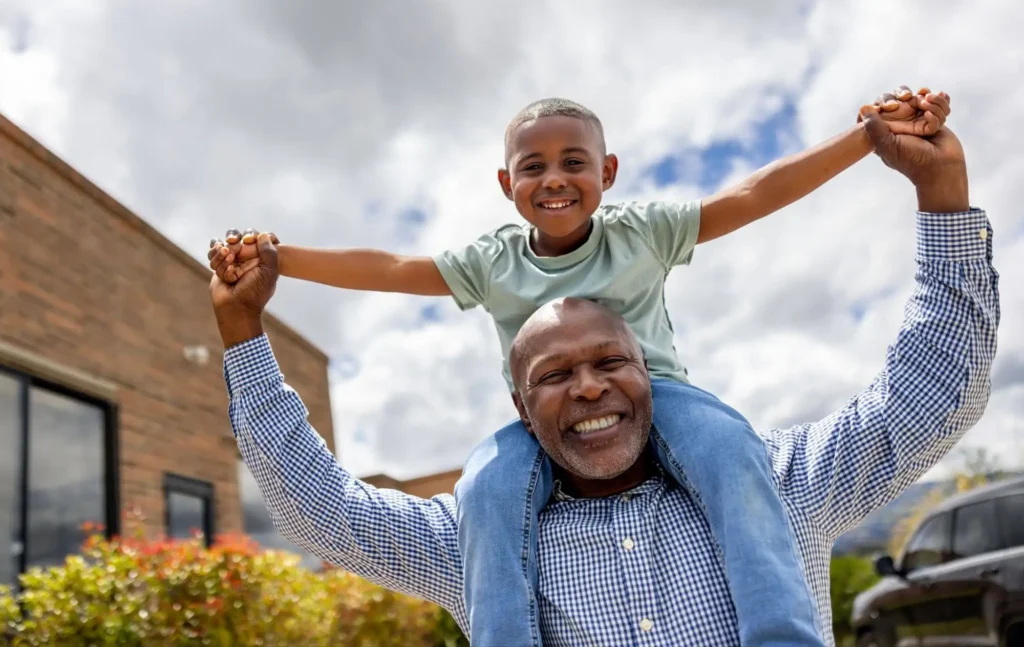 An elderly man carrying grandchild on shoulder while holding hands and smiling from his home at Village Shalom, offering senior living in Overland Park, KS.